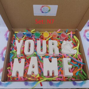 May include: A craft kit in a cardboard box, featuring the words "YOUR NAME" in large, white block letters, a white unicorn figurine, and small paint pots. The box is filled with colorful shredded paper. The text "Set: N7" is visible.