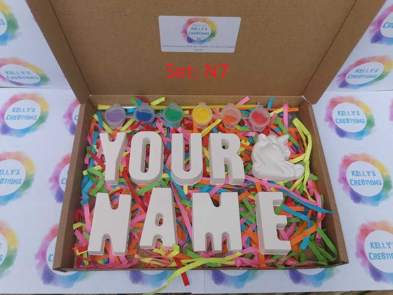 May include: A craft kit in a cardboard box, featuring the words "YOUR NAME" in large, white block letters, a white unicorn figurine, and small paint pots. The box is filled with colorful shredded paper. The text "Set: N7" is visible.