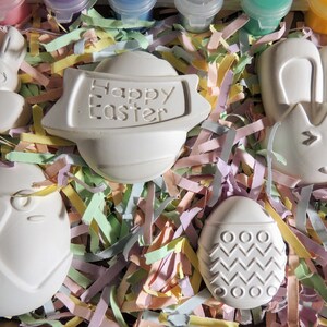 May include: A set of five white ceramic Easter-themed ornaments, including a bunny, an egg with a zigzag pattern, an egg with a bird design, a bunny with ears, and a round ornament with the text "Happy Easter". The ornaments are nestled in pastel-colored shredded paper.