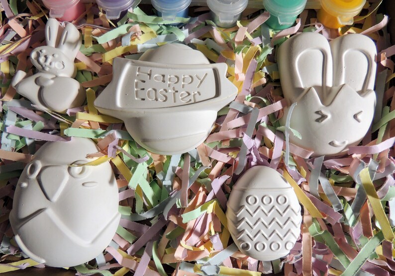 May include: A set of five white ceramic Easter-themed ornaments, including a bunny, an egg with a zigzag pattern, an egg with a bird design, a bunny with ears, and a round ornament with the text "Happy Easter". The ornaments are nestled in pastel-colored shredded paper.
