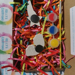 May include: A craft kit with two white ceramic shapes, a star and a heart, with black magnetic circles attached. The kit includes three small pots of paint, red, blue, and yellow, and a small paintbrush. The kit is packaged in a cardboard box with colorful shredded paper.