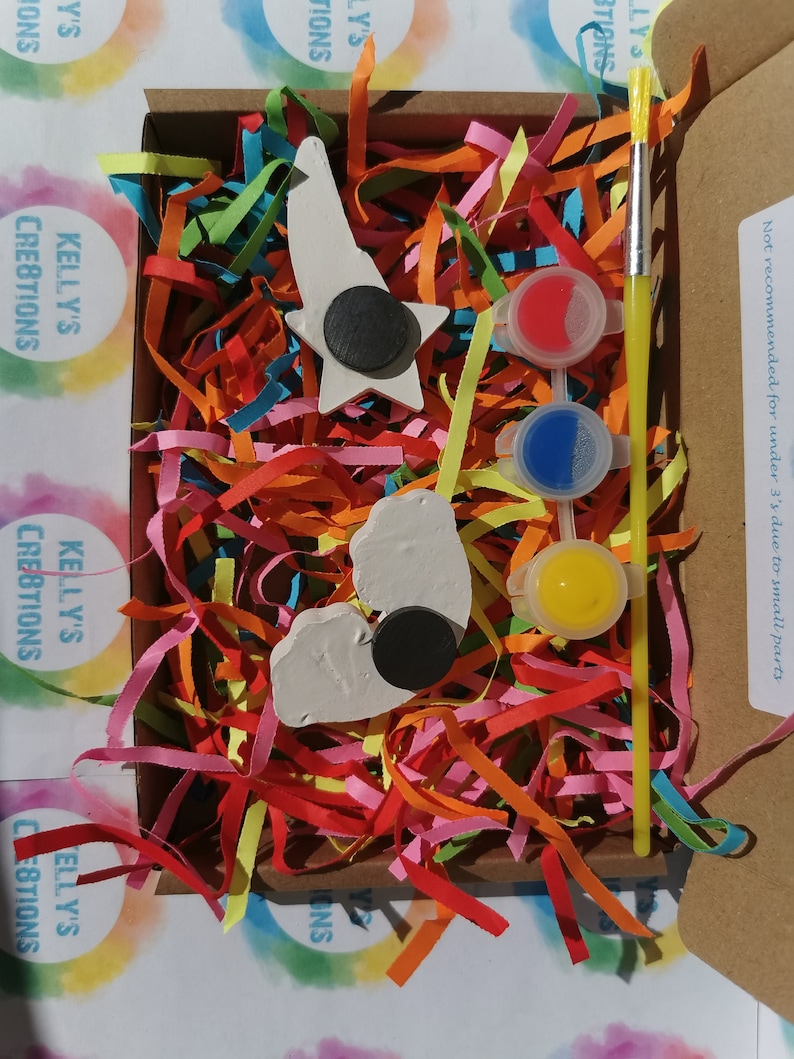 May include: A craft kit with two white ceramic shapes, a star and a heart, with black magnetic circles attached. The kit includes three small pots of paint, red, blue, and yellow, and a small paintbrush. The kit is packaged in a cardboard box with colorful shredded paper.