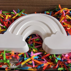 May include: A white ceramic rainbow with clouds on either side, sitting in a box filled with colorful shredded paper.
