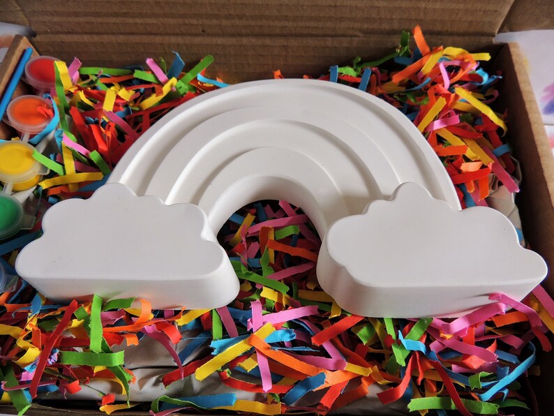 May include: A white ceramic rainbow with clouds on either side, sitting in a box filled with colorful shredded paper.