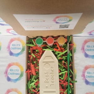 May include: A white ceramic pencil-shaped ornament with the text "Best Teacher" and a red apple design. The ornament is in a box with green and red shredded paper, three small paint pots, and a paintbrush.