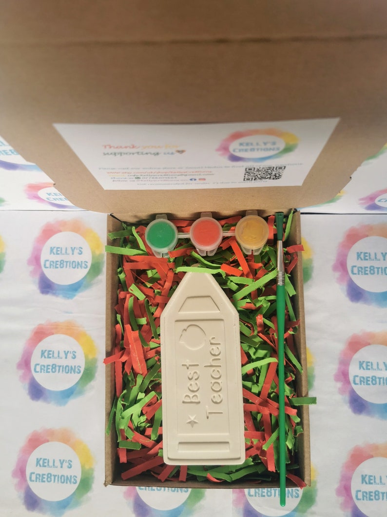 May include: A white ceramic pencil-shaped ornament with the text "Best Teacher" and a red apple design. The ornament is in a box with green and red shredded paper, three small paint pots, and a paintbrush.