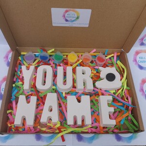 May include: A craft kit in a cardboard box, featuring the words "YOUR NAME" in white block letters, a sewing machine shape, and small paint pots. The box is filled with colorful shredded paper.