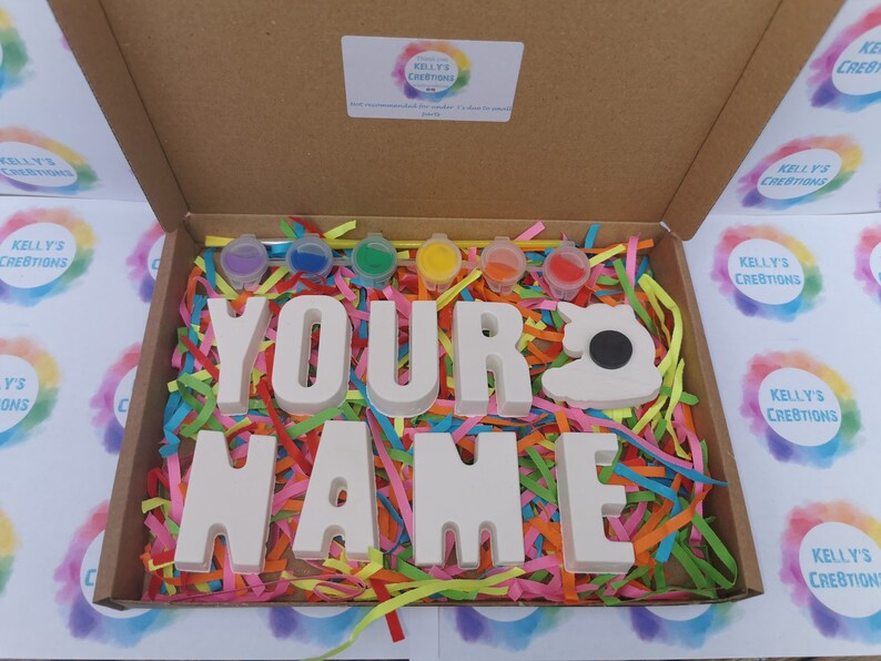 May include: A craft kit in a cardboard box, featuring the words "YOUR NAME" in white block letters, a sewing machine shape, and small paint pots. The box is filled with colorful shredded paper.