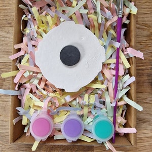 May include: A craft kit with a white ceramic blank, three small paint pots in pink, purple, and teal, and a paintbrush. The blank has a black circle in the center.