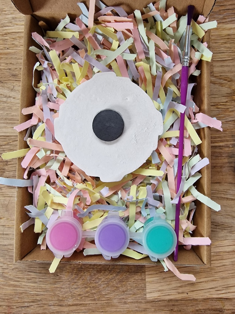 May include: A craft kit with a white ceramic blank, three small paint pots in pink, purple, and teal, and a paintbrush. The blank has a black circle in the center.