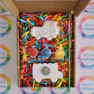 Puede incluir: Un kit de manualidades en una caja de cart&oacute;n, con una pieza de cer&aacute;mica blanca en forma de cami&oacute;n, botes de pintura roja, azul y amarilla, y un pincel verde. La caja est&aacute; llena de papel triturado de colores. Tambi&eacute;n se incluye una segunda pieza en forma de cami&oacute;n con un im&aacute;n.