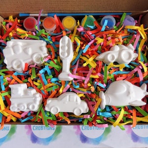 May include: A set of six white ceramic figurines of different vehicles, including a car, truck, train, airplane, and traffic light, in a box with colorful shredded paper and paint pots.