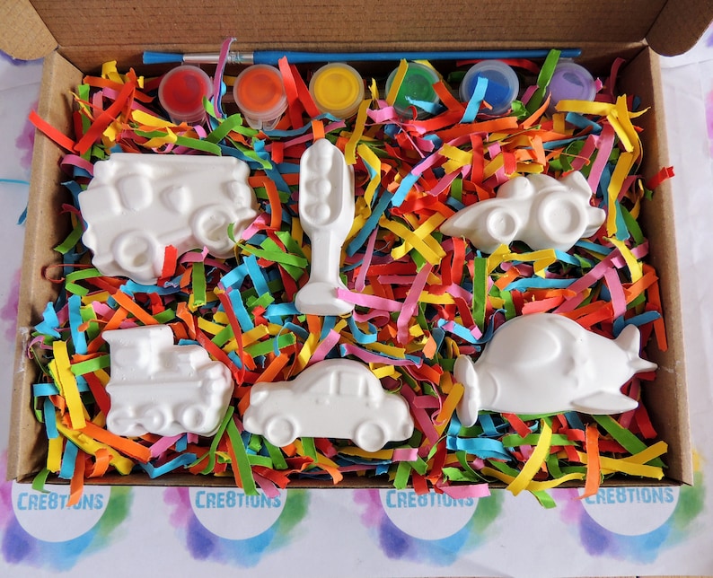May include: A set of six white ceramic figurines of different vehicles, including a car, truck, train, airplane, and traffic light, in a box with colorful shredded paper and paint pots.