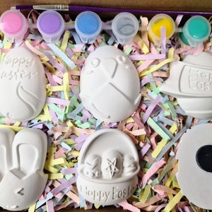 May include: A set of six white ceramic Easter-themed ornaments in a box with pastel-coloured shredded paper. The ornaments include a bunny, a basket with eggs, an egg with the words "Happy Easter", an egg with a cross, an egg with a chick, and a round ornament with a magnet.