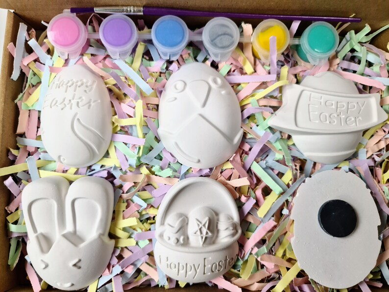 May include: A set of six white ceramic Easter-themed ornaments in a box with pastel-coloured shredded paper. The ornaments include a bunny, a basket with eggs, an egg with the words "Happy Easter", an egg with a cross, an egg with a chick, and a round ornament with a magnet.