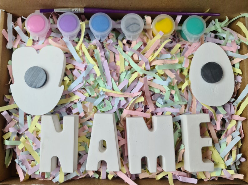 May include: A craft kit for painting a name with white ceramic letters and two white ceramic shapes, one shaped like an egg and the other like a flower. The kit includes six small paint pots with lids in pink, purple, blue, gray, yellow, and green. A small paintbrush is included.