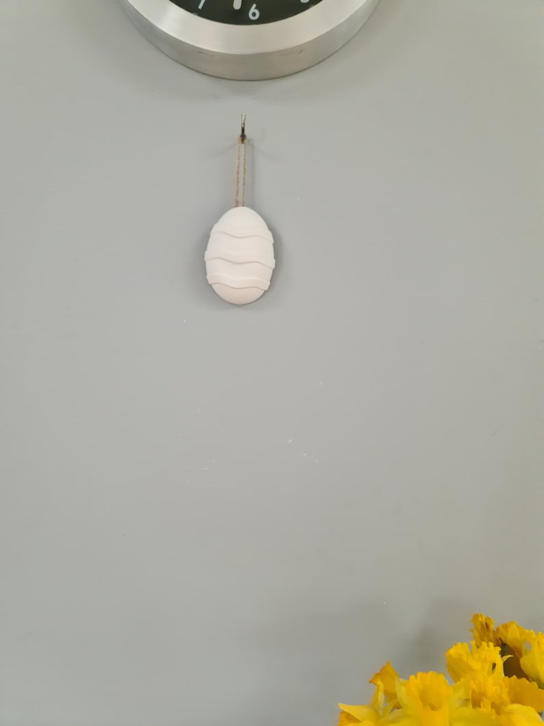 Puede incluir: Un adorno de huevo de Pascua de cer&aacute;mica blanca con un dise&ntilde;o ondulado cuelga de un gancho en una pared gris claro.