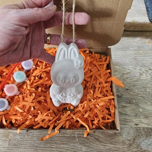 May include: A white ceramic Halloween bunny ornament with a skull on its head, hanging from a twine string. The ornament is inside a cardboard box filled with orange paper shreds. Paint pots and brushes are visible.