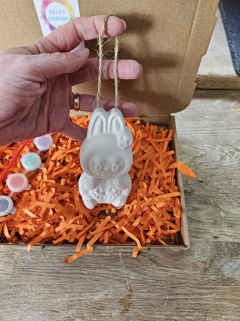 May include: A white ceramic Halloween bunny ornament with a skull on its head, hanging from a twine string. The ornament is inside a cardboard box filled with orange paper shreds. Paint pots and brushes are visible.