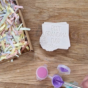 May include: A white ceramic tile with the text "Best Grandma Ever" in a black frame. The tile is surrounded by a floral design. Three small paint pots with pink, purple, and turquoise paint are on a wooden surface. A paintbrush is dipping into the purple paint.