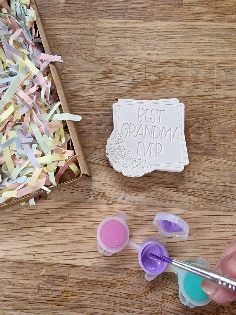 May include: A white ceramic tile with the text "Best Grandma Ever" in a black frame. The tile is surrounded by a floral design. Three small paint pots with pink, purple, and turquoise paint are on a wooden surface. A paintbrush is dipping into the purple paint.