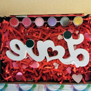 May include: A white wooden sign with the word "love" painted on it. The sign is surrounded by red and pink tissue paper and comes with a set of six paint pots in various colours.