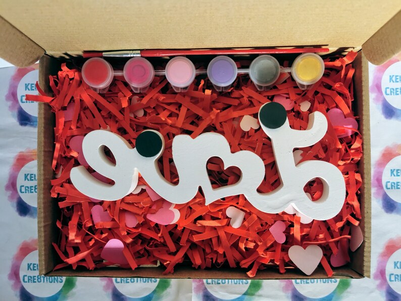 May include: A white wooden sign with the word "love" painted on it. The sign is surrounded by red and pink tissue paper and comes with a set of six paint pots in various colours.
