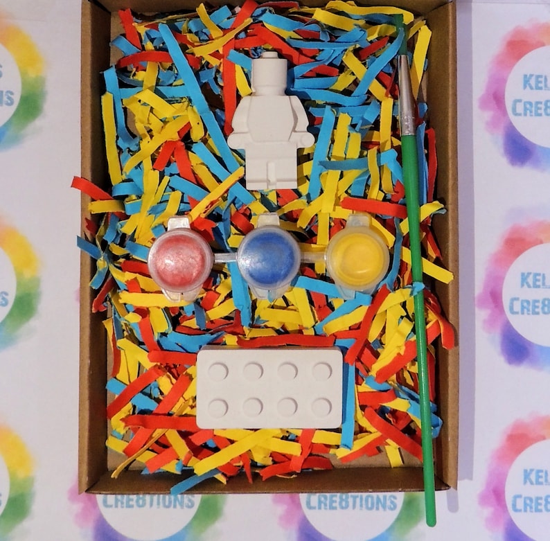 Puede incluir: Una figura de Lego de pl&aacute;stico blanco, tres peque&ntilde;os botes de pintura roja, azul y amarilla, una base de ladrillo Lego de pl&aacute;stico blanco y un pincel verde en una caja de cart&oacute;n llena de papel triturado rojo, amarillo y azul.
