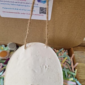 May include: A white, egg-shaped craft kit with a scalloped bottom edge, suspended by a jute string. The kit is in a cardboard box filled with colorful paper shreds. The kit includes paint and instructions.