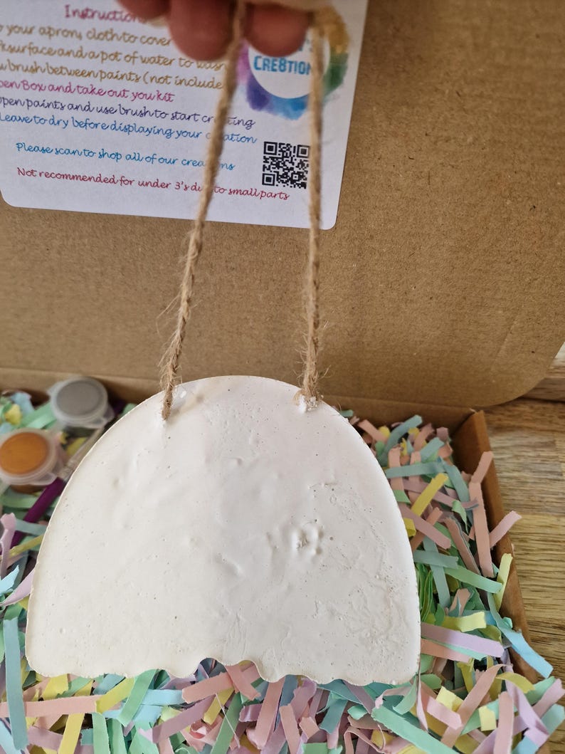 May include: A white, egg-shaped craft kit with a scalloped bottom edge, suspended by a jute string. The kit is in a cardboard box filled with colorful paper shreds. The kit includes paint and instructions.