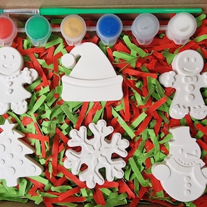 May include: A Christmas-themed paint-your-own craft kit with six white ceramic ornaments: a gingerbread person, a Christmas tree, a snowflake, a Santa hat, a snowman, and another gingerbread person. The kit includes six paint pots in red, green, yellow, blue, white, and silver.