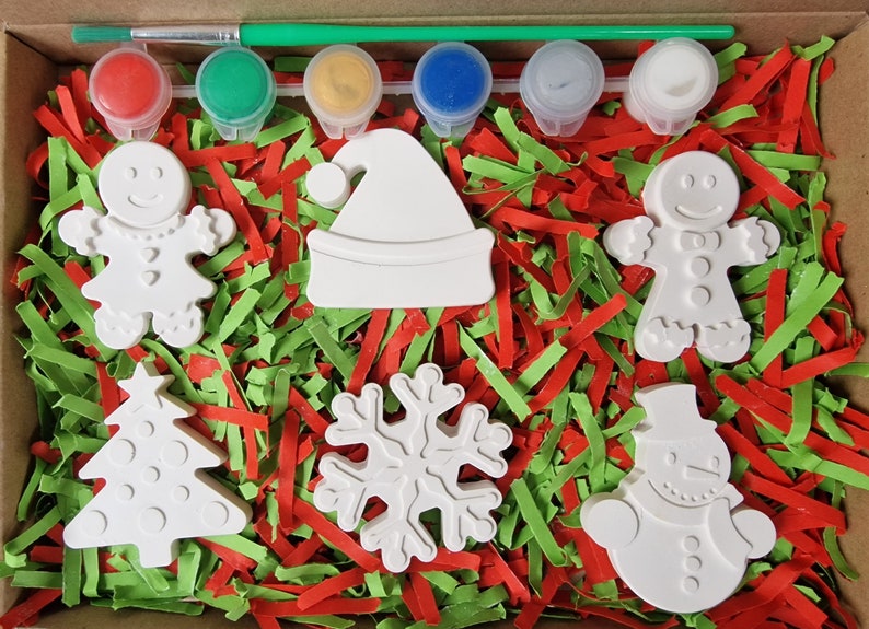 May include: A Christmas-themed paint-your-own craft kit with six white ceramic ornaments: a gingerbread person, a Christmas tree, a snowflake, a Santa hat, a snowman, and another gingerbread person. The kit includes six paint pots in red, green, yellow, blue, white, and silver.