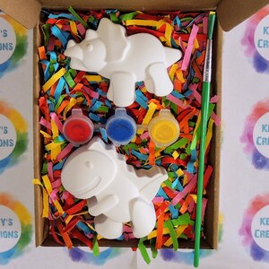 May include: A craft kit with two white ceramic dinosaur figurines, red, blue, and yellow paint pots, and a green paintbrush. The dinosaurs are in a box filled with colorful shredded paper.