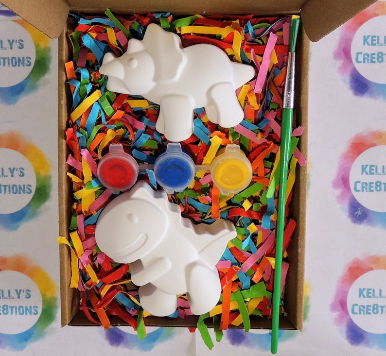 May include: A craft kit with two white ceramic dinosaur figurines, red, blue, and yellow paint pots, and a green paintbrush. The dinosaurs are in a box filled with colorful shredded paper.