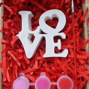 May include: A white wooden "LOVE" sign with a heart cutout in the "O" is surrounded by red shredded paper. Three small paint pots with pink and red paint are included, along with a paintbrush.