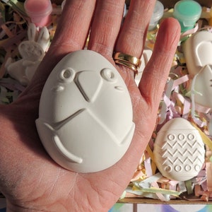 May include: A set of four white ceramic Easter-themed ornaments, including a bunny, an egg with a bird face, and two eggs with geometric designs. The ornaments are in a box with colorful shredded paper.