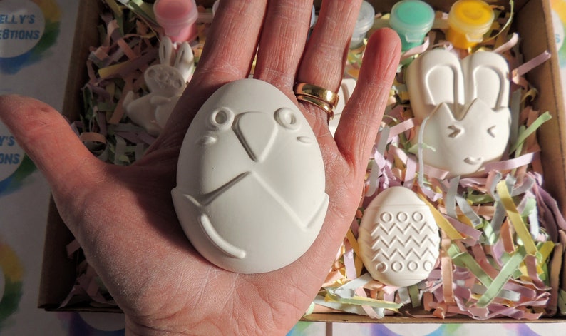 May include: A set of four white ceramic Easter-themed ornaments, including a bunny, an egg with a bird face, and two eggs with geometric designs. The ornaments are in a box with colorful shredded paper.