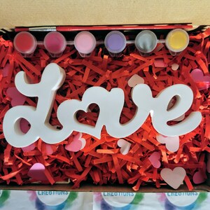 May include: A white wooden "Love" sign with a heart cutout in the "O" is nestled in red and pink shredded paper. Five small paint pots with red, pink, purple, grey, and yellow paint are included, along with a paintbrush.