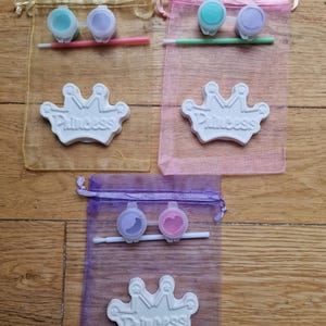May include: Three organza bags with paint and a brush for painting a white ceramic crown with the word "Princess" on it.