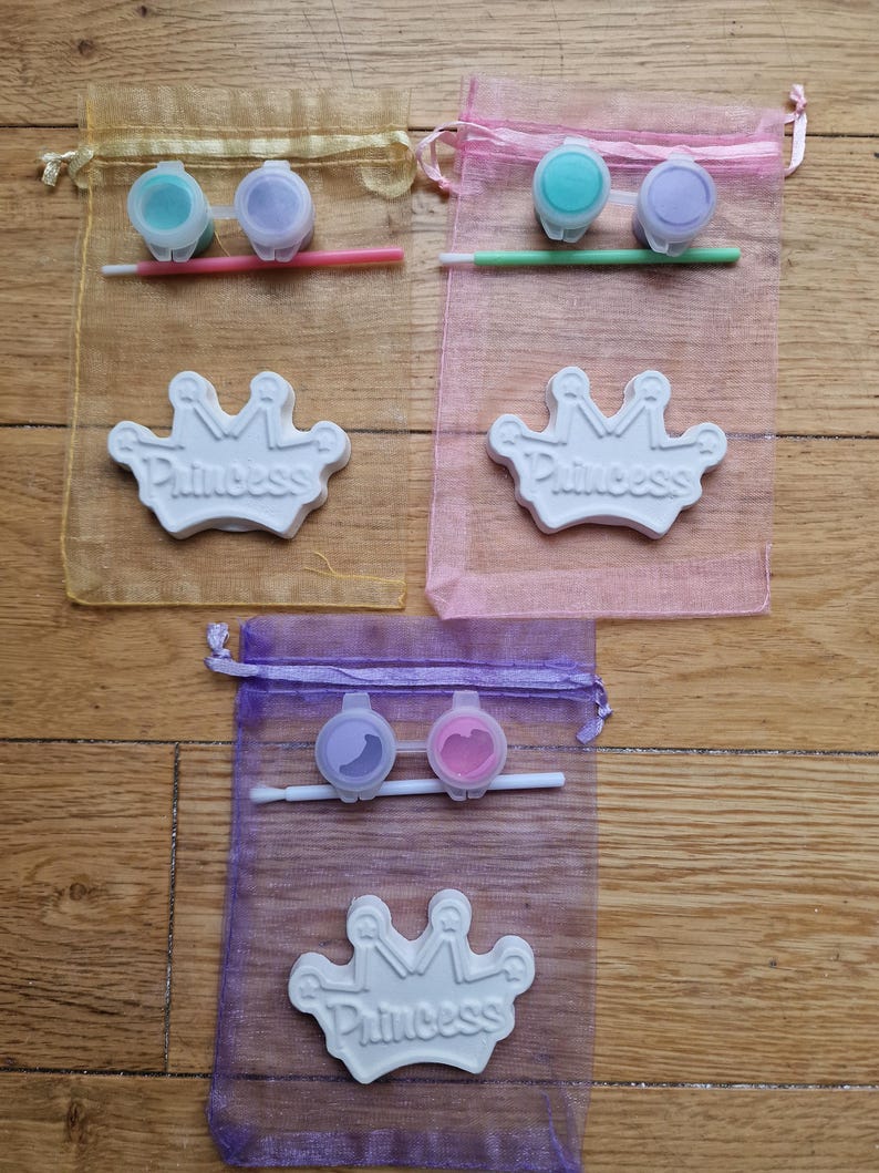 May include: Three organza bags with paint and a brush for painting a white ceramic crown with the word "Princess" on it.