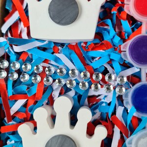 May include: Two white ceramic crown shapes with grey circles in the centre. The crowns are surrounded by blue, red and white shredded paper.