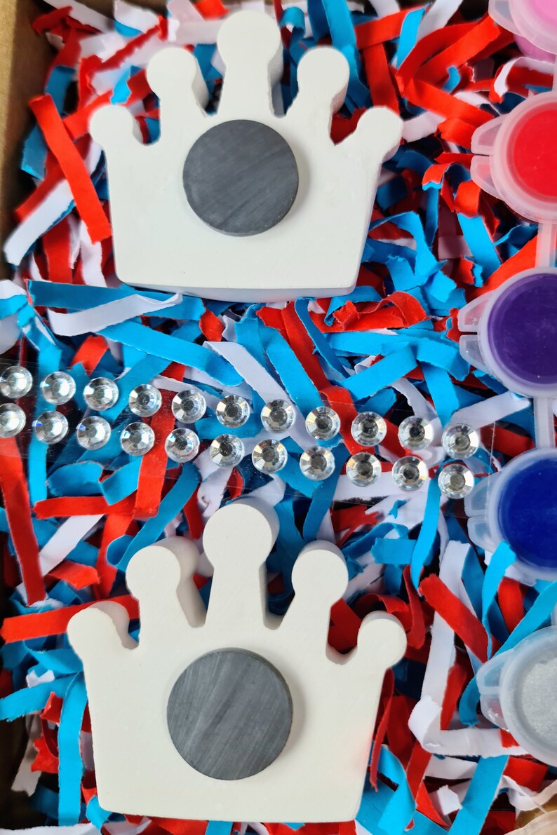May include: Two white ceramic crown shapes with grey circles in the centre. The crowns are surrounded by blue, red and white shredded paper.