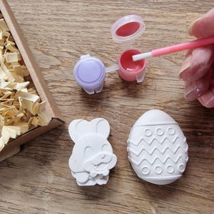 May include: A craft kit with two white ceramic figurines, one shaped like an Easter egg with a zig-zag pattern and the other shaped like a bunny holding an egg. The kit includes two small pots of paint, one purple and one pink, and a paintbrush.