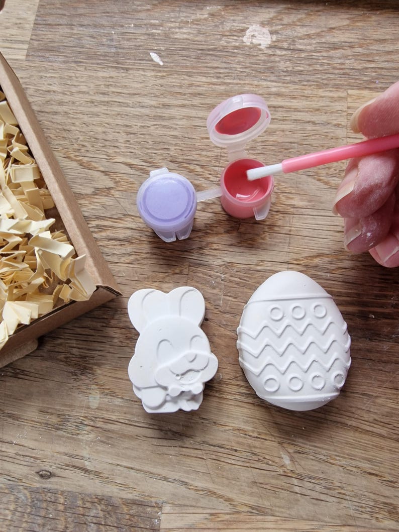 May include: A craft kit with two white ceramic figurines, one shaped like an Easter egg with a zig-zag pattern and the other shaped like a bunny holding an egg. The kit includes two small pots of paint, one purple and one pink, and a paintbrush.
