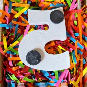 Puede incluir: Un n&uacute;mero cinco de madera blanco con dos c&iacute;rculos negros, botes de pintura en rojo, azul y amarillo, y un pincel en una caja llena de papel triturado de colores.