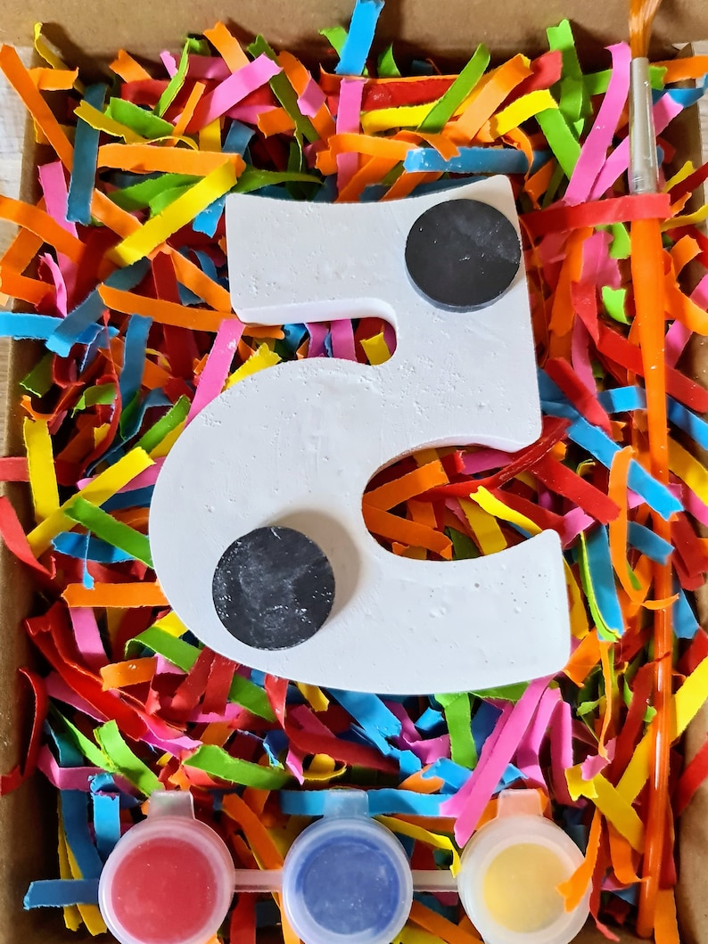 Puede incluir: Un n&uacute;mero cinco de madera blanco con dos c&iacute;rculos negros, botes de pintura en rojo, azul y amarillo, y un pincel en una caja llena de papel triturado de colores.