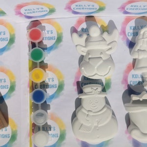 May include: A craft kit with unpainted Christmas-themed ceramic figurines, including a snowman, reindeer, Santa Claus, and a sleigh. The kit includes paint pots in red, green, yellow, and blue, along with shredded paper for packaging. The kit is labeled "Kelly's Creations".