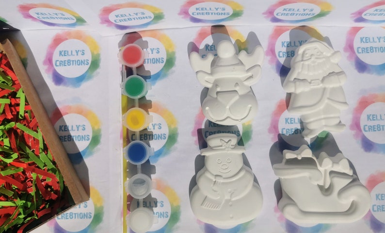 May include: A craft kit with unpainted Christmas-themed ceramic figurines, including a snowman, reindeer, Santa Claus, and a sleigh. The kit includes paint pots in red, green, yellow, and blue, along with shredded paper for packaging. The kit is labeled "Kelly's Creations".