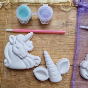 Puede incluir: Un kit de manualidades con dos figuritas de unicornio de cer&aacute;mica blanca, una con un cuerno y orejas, y la otra con una cabeza y melena completas. El kit incluye dos peque&ntilde;os botes de pintura, uno turquesa y otro morado, y un pincel rosa.
