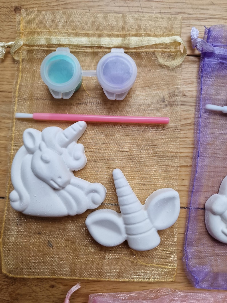 Puede incluir: Un kit de manualidades con dos figuritas de unicornio de cer&aacute;mica blanca, una con un cuerno y orejas, y la otra con una cabeza y melena completas. El kit incluye dos peque&ntilde;os botes de pintura, uno turquesa y otro morado, y un pincel rosa.